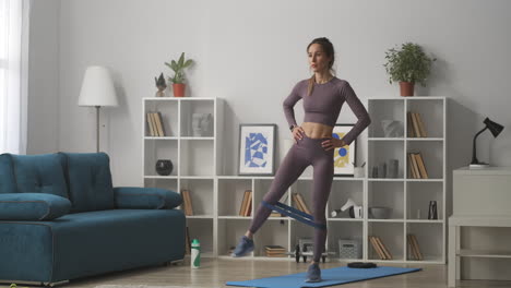 aerobics-and-gymnastics-training-at-home-woman-is-moving-legs-standing-in-living-room-sporty-female-body-dressed-trendy-sportswear-healthy-lifestyle
